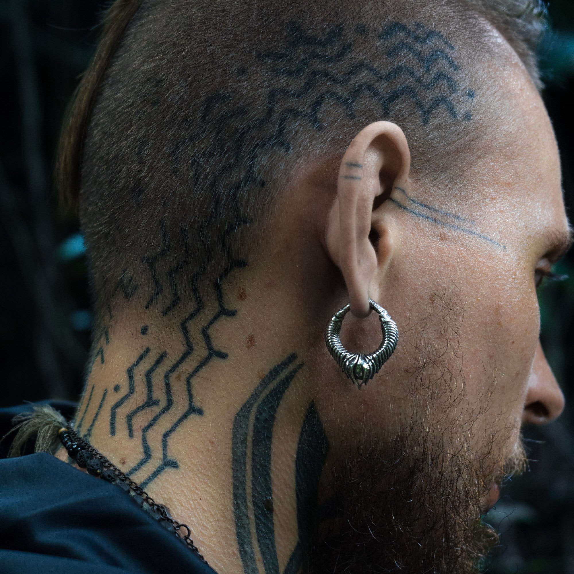 A close-up side profile of a person with a partially shaved head and intricate, zigzag tattoos on the scalp and neck. The person has a beard and is wearing Kaya Movement's MARVDASHT Small Biomechanical Clicker Ear Weights in 925 Silver, designed in a 2 gauge size that add subtle sci-fi vibes. The background is blurred and dark.